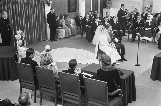Schwarzes und weißes Foto einer Hochzeitszeremonie mit sitzenden Gästen, einem Mann, der in ein Mikrofon spricht, einer Frau mit einem Strauß Blumen, einem Podium mit Mikrofon, Blumenschmuck, einem Vorhang, einem gerahmten Foto an der Wand und einer Tür im Hintergrund.