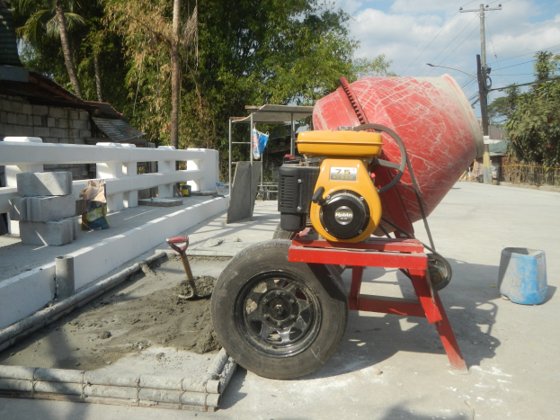Betonmischer auf einem Zementlaster umgeben von Baustoffen und einer Wohngegend bei bewölktem Himmel.