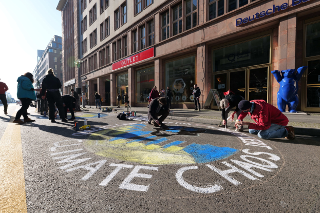 Eine Gruppe von Menschen sitzt vor einem Gebäude mit Fenstern und Namensschildern während einer Klimaprotest in Berlin auf dem Boden, umgeben von Flaschen und anderen Gegenständen, mit Bäumen und einem klaren blauen Himmel im Hintergrund.