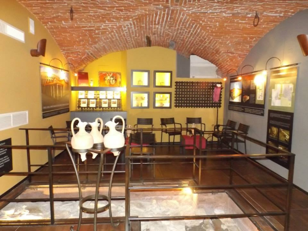 Ein geräumiger Museumsraum mit mehreren Tischen, Stühlen, Displaytafeln, gerahmten Kunstwerken an den Wänden, Vasen, Leuchten und verschiedenen anderen Gegenständen, die für die Ausstellung angeordnet sind.