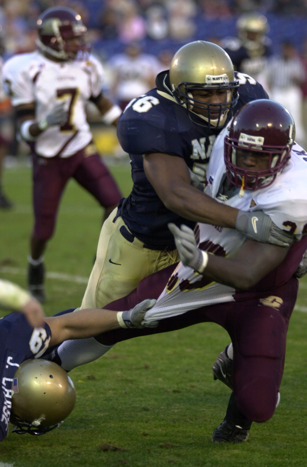 Zwei Fußballspieler in Sportkleidung und Helmen, einer hält den anderen fest, mit zusätzlichen Spielern und Zuschauern im Hintergrund.